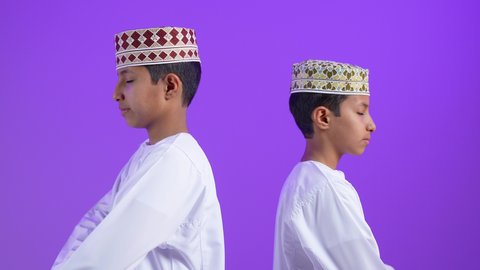 تعابير الفرح والسرور، الاهتمام بالأناقة والمظهر الخارجي، بورترية لطفلان توأمان عربيان خليجيان عمانيان يرتديان الدشداشة و الكمه يقفان بأيدي متقاطعة ينظران إلى الكاميرا بإيماءات الثقة بالنفس، خلفية بنفسجية