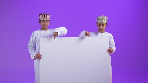 لوح إعلاني فارغ، موك اب أبيض، بورترية لطفلان توأمان عربيان خليجيان عمانيان يرتديان الدشداشة و الكمه يحملان بيدهما لوحه بيضاء و يقومان بالاشارة اليها , خلفية بنفسجية