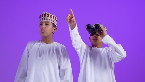حب المغامرة والاستكشاف، رؤية الفرص والتحديات المحتملة، الاشارة الي شي ما، بورتريه لطفل عربي خليجي عماني يرتدي الدشداشة و الكمة ينظر من خلال المنظار ثنائي العينين برفقة توأمه، رؤية الاشياء البعيدة عبر التلسكوب، ايماءات الاندهاش و التعجب ، خلفية بنفسجية