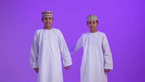 ايماءات يدين على شكل قلب، اظهار مشاعر الحب والشغف، مفهوم المحبة والمودة، بورتريه لطفلان توأمان عربيان خليجيان عمانيان يرتديان الدشداشة و الكمه ينظران إلى الكاميرا بتعابير السرور والسعادة، خلفية بنفسجية