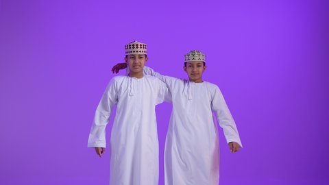 إشارات وحركات يدين تدل على هذا المكان (هنا)، تحديد موقع معين، بورتريه لطفلان توأمان عربيان خليجيان عمانيان يرتديان الدشداشة و الكمه يشيران بأصابعهما نحو الأعلى، خلفية بنفسجية