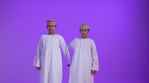 تحديد موقع معين ، إشارات وحركات يدين تدل على هذا المكان (هنا)، بورتريه لطفلان توأمان عربيان خليجيان عمانيان يرتديان الدشداشة و الكمه يشيران بأصابعهما نحو الأعلى، خلفية بنفسجية