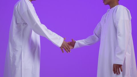Building strong social relationships in Omani society, the concept of forgiveness and tolerance, greeting and peace, close-up portrait of two Omani Gulf Arab twin children wearing traditional dress shaking hands, purple background