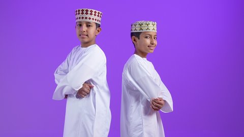 الاهتمام بالأناقة والمظهر الخارجي، بورتريه لطفلان توأمان عربيان خليجيان عمانيان يرتديان الدشداشة و الكمه يقفان بأيدي متقاطعة ينظران إلى الكاميرا بإيماءات الثقة بالنفس، تعابير الفرح والسرور، خلفية بنفسجية
