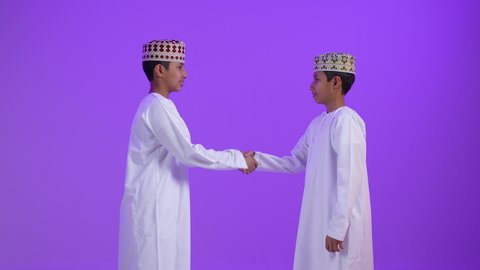 مفهوم الصفح والتسامح، إلقاء السلام والتحية, بورترية لطفلان توأمان عربيان خليجيان عمانيان يرتديان الدشداشة و الكمه يتصافحان سوياً, بناء العلاقات الاجتماعية المتينة في المجتمع العماني ، خلفية بنفسجية