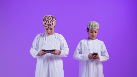 الالعاب المسليه وقت الفراغ، استخدام الأجهزة والتقنيات الحديثة، التواصل و الدردشة مع الاصدقاء، بورتريه لطفل عربي خليجي عماني يرتدي الدشداشة و الكمه يمسك بيده الهاتف المحمول برفقه توأمه، ايماءات السعاده و السرور، خلفية بنفسجية