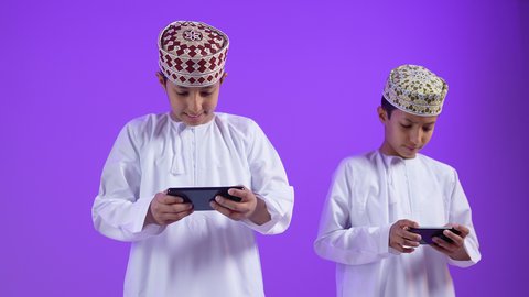 Using modern devices and technologies, communication and chatting with friends, portrait of an Arab Gulf Omani child wearing a dishdasha and a sleeve, holding a mobile phone with his twin, entertaining games in free time, gestures of happiness and pleasure, purple background
