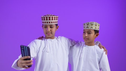 ايماءات وتعابير البهجة والسرور، استعمال الأجهزة التقنية الحديثة في اخذ صور تذكارية، بورتريه لطفل عربي خليجي عماني يرتدي الدشداشة و الكمه يقوم بإلتقاط صورة سيلفي برفقة توأمه باستخدام الهاتف المحمول، خلفية  بنفسجية