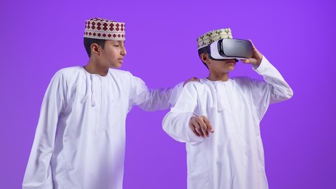المتعة والتسلية بالمشاهدة عبر نظارة الواقع الافتراضي, بورترية لطفل عربي خليجي عماني يرتدي الدشداشة و الكمه يضع نظارات ثلاثية الأبعاد بتعابير الاندماج والحماس برفقة توأمه، محاكاة العالم الخيالي، استخدام جهاز تقني حديث ومتطور، خلفية بنفسجية