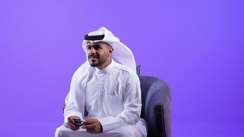 تعابير التفاعل والاندماج مع ألعاب الفيديو، شاب سعودي يقضي وقت ممتع ومسلي، رجل عربي خليجي كويتي يرتدي الثوب التقليدي يجلس على كرسي مريح يمسك بيديه عصا التحكم، استخدام الأجهزة والتقنيات الإلكترونية الحديثة