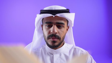 مفهوم الثقافة والتعلم الذاتي, الاستمتاع بالأنشطة الترفيهية المفيدة في وقت الفراغ, قراءة القصص والروايات,تصوير مقرب لشاب عربي خليجي كويتي يرتدي الثوب التقليدي يجلس على كرسي مريح يحمل كتاب وينظر اليه بايماءات الاندماج ، رجل سعودي يمارس هواية المطالعة