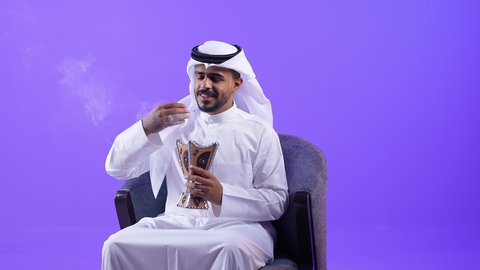 العادات و التقاليد العريقه في الكويت, النظر إلى الكاميرا بايماءات الفرح والسرور, رجل سعودي يبخر بخشب العود في الاعياد والمناسبات، شاب عربي خليجي كويتي يرتدي الثوب التقليدي والغتره يجلس على كرسي مريح يقوم بالتطيب و التعطر من البخور( المبخرة )