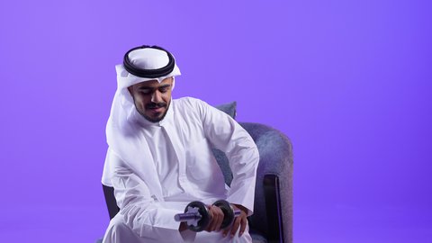 ممارسة التمارين الرياضية ورفع الأثقال مع الدمبل ،مفهوم اللياقة الجسدية والرشاقة البدنية، رجل سعودي يقوم بتقوية عضلات الجسم و بنائها ، شاب عربي خليجي كويتي يرتدي الثوب التقليدي يجلس على الكرسي المريح يحمل بيد الأوزان الحديدية الثقيلة يشير إليها