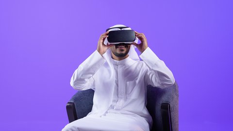 المتعة والتسلية بالمشاهدة عبر نظارة الواقع الافتراضي, شاب عربي خليجي كويتي يرتدي الثوب التقليدي والغترة يجلس على كرسي يضع نظارات ثلاثية الأبعاد يقوم برفع كف يده للاعلى بتعابير الاندماج , رجل سعودي يشير بايماءات التوقف عن فعل امر ما