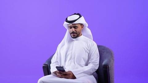 تصفح مواقع وتطبيقات الانترنت، النظر إلى الهاتف المحمول بإيماءات الاندماج، الدردشة والتحدث مع الأصدقاء، شاب عربي خليجي كويتي مبتسم يرتدي الثوب التقليدي والغتره يمسك الجوال بيده، رجل سعودي يستخدم التقنيات والتكنولوجيا الحديثة بالحياة اليومية