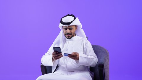 سهولة الشراء والدفع عن طريق بطاقة الإئتمان البنكية، استخدام الأجهزة والتقنيات الحديثة, شاب عربي خليجي كويتي يجلس على كرسي مريح يرتدي الثوب التقليدي والغتره يمسك بيده بطاقة الصراف و الهاتف المحمول, رجل سعودي يتسوق عبر الانترنت