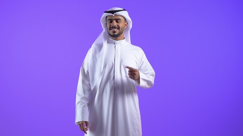 شاب عربي خليجي كويتي مبتسم يرتدي الثوب التقليدي والغترة يقف ويشير بإصبعيه السبابة جانبا إلى شيء محدد بتعابير هذا المكان هنا، رجل سعودي يحدد موقع معين ، النظر إلى الكاميرا بإيماءات الفرح والسرور