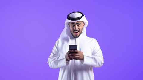 النظر إلى الهاتف المحمول بإيماءات الاندماج،  تصفح مواقع وتطبيقات الانترنت، الدردشة والتحدث مع الأصدقاء، شاب عربي خليجي كويتي مبتسم يرتدي الثوب التقليدي والغتره يمسك الجوال بيده، رجل سعودي يستخدم التقنيات والتكنولوجيا الحديثة بالحياة اليومية
