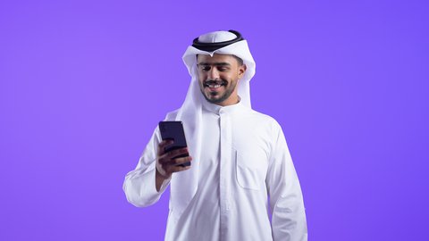 تصفح مواقع وتطبيقات الانترنت، النظر إلى الهاتف المحمول بإيماءات الاندماج، الدردشة والتحدث مع الأصدقاء، شاب عربي خليجي كويتي مبتسم يرتدي الثوب التقليدي والغتره يمسك الجوال بيده، رجل سعودي يستخدم التقنيات والتكنولوجيا الحديثة بالحياة اليومية