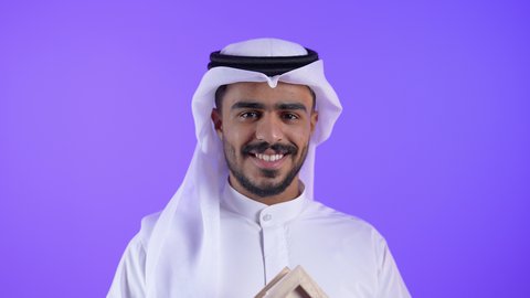 شاب عربي خليجي كويتي يرتدي الثوب التقليدي والغترة يحمل بيده مجسم خشبي لبيت ، ايماءات السعادة والسرور ،رجل سعودي ينظر الى الكاميرا بايماءات البهجة لامتلاك سكن حديث