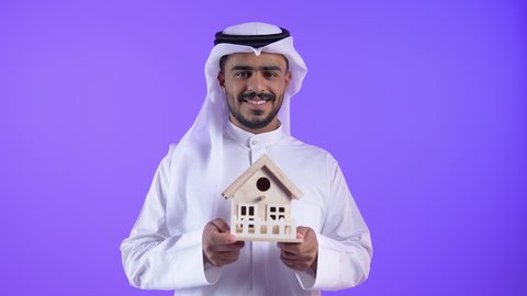ايماءات السعادة والسرور ، شاب عربي خليجي كويتي يرتدي الثوب التقليدي والغترة يحمل بيده مجسم خشبي لبيت ، رجل سعودي ينظر الى الكاميرا بايماءات البهجة لامتلاك سكن حديث
