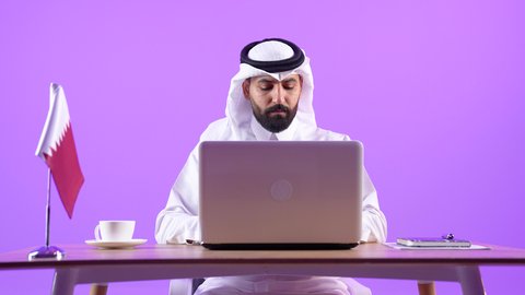 Focus, dedication and hard work, use of modern technologies in administrative and office work, employment in Qatari offices and companies, portrait of a Qatari Gulf Arab man wearing traditional thobe and ghutra sitting behind a desk with the Qatari flag next to him and a laptop in front of him, purple background
