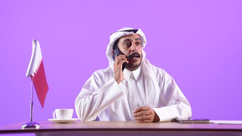 استخدام الهاتف المحمول في العمل، شاب سعودي يقوم بالرد والإجابة على الأسئلة والاستفسارات والمكالمات، بورتريه مقرب لرجل عربي خليجي قطري يرتدي الثوب التقليدي والغترة يجلس خلف المكتب أمامه الحاسوب المحمول وعلم قطر، خلفية بنفسجية