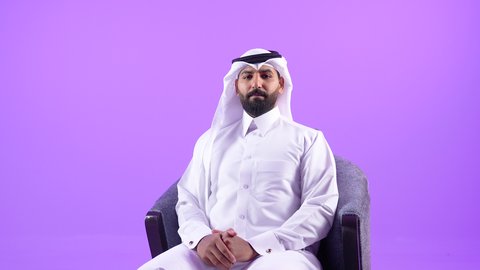 مفهوم الوسامة والرجولة والقوة، شاب سعودي يقوم بالاهتمام بالأناقة والمظهر الخارجي، بورتريه لرجل عربي خليجي قطري يرتدي الثوب التقليدي والغترة يجلس على كرسي مريح ينظر الى الكاميرا بإيماءات السرور والثقة في النفس، خلفية بنفسجية