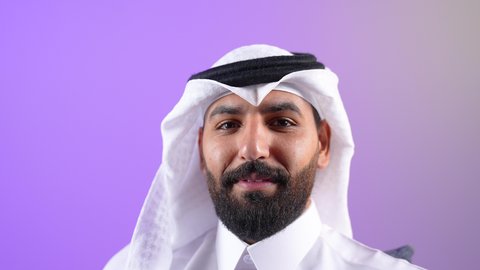 النظر الى الكاميرا بإيماءات السرور والثقة في النفس، مفهوم الوسامة والرجولة والقوة، شاب سعودي يقوم بالاهتمام بالأناقة والمظهر الخارجي، بورتريه مقرب لرجل عربي خليجي قطري يرتدي الثوب التقليدي والغترة يجلس على كرسي مريح، خلفية بنفسجية