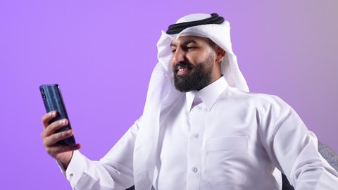 التواصل مع الافراد عبر الهاتف المحمول، ايماءات السعادة و السرور، دردشة ومحادثات وتعارف، بورتريه مقرب لرجل عربي خليجي قطري يرتدي الثوب التقليدي والغترة يجلس على كنبة مريحة يستعمل الجوال، شاب سعودي يقوم باجراء مكالمة فيديو, استخدام التقنيات والأجهزة الحديثة، خلفية بنفسجية
