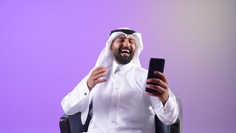 Gestures of happiness and pleasure, communicating with people via mobile phone, chatting, conversations and getting to know each other, portrait of a Qatari Gulf Arab man wearing traditional thobe and ghutra sitting on a comfortable sofa using a mobile phone, a Saudi young man making a video call, using modern technologies and devices, purple background