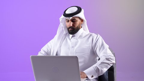 مفهوم التوظيف أونلاين، رجل سعودي يقوم بتصفح مواقع التواصل الاجتماعي بواسطة الحاسب المحمول، العمل عن بعد عبر الانترنت، دمج التكنولوجيا في الحياة العملية, بورتريه لرجل عربي خليجي قطري يرتدي الثوب التقليدي والغترة يجلس على كرسي مريح يقوم باستخدام جهاز اللابتوب، خلفية بنقسجية