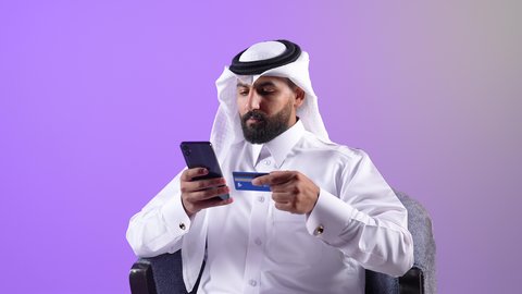الدفع الالكتروني بالاعتماد على البطاقة البنكية،شاب سعودي يقوم باستخدام التقنيات الحديثة والتكنولوجيا, بورتريه مقرب لرجل عربي خليجي قطري يرتدي الثوب التقليدي والغترة يعمل على جهاز الهاتف المحمول، مفهوم التسوق أونلاين، عمليات التبضع والشراء عبر تطبيقات الانترنت، خلفية بنفسجية