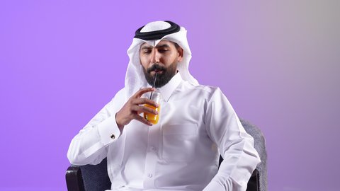الحصول على كأس من العصير البارد والمنعش، شاب سعودي يقوم بالجلوس ومحاولة الراحة والاستمتاع بأوقات الفراغ، بورتريه مقرب لشاب عربي خليجي قطري يرتدي الثوب التقليدي والغترة يجلس على كرسي مريح يمسك بيده كوب من المشروب الصيفي المفضل، خلفية بنفسجية