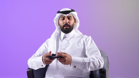 Gestures of happiness and enjoyment using modern technologies, a Saudi young man interacting and merging with entertainment video games, a portrait of a smiling Qatari Gulf Arab man wearing a thobe and ghutra sitting on a comfortable chair holding a joystick with expressions of integration and pleasure, purple background