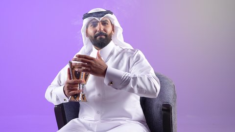 التبخر في الأعياد والمناسبات السعيدة، اتباع العادات والتقاليد العربية الأصيلة، شاب سعودي يقوم بالتبخير واستخدام العطور والمطيبات، بورتريه  لرجل عربي خليجي قطري يرتدي الثوب التقليدي والغترة يجلس على كرسي ويمسك بيده المبخرة، خلفية بنفسجية