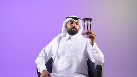 Business management and schedule organization concept, Saudi young man feeling aware of the value of precious moments, portrait of a Qatari Gulf Arab man wearing traditional thobe and ghutra holding a glass sand timer (bankam) in his hand and monitoring the sand timing, purple background