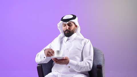 Relaxing and having a hot drink, trying to get some rest, portrait of a Qatari Gulf Arab man wearing traditional thobe and ghutra sitting on a chair holding a cup of his favorite drink, a Saudi young man enjoying a cup of hot tea, purple background