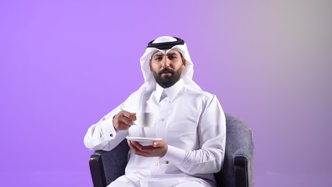 محاولة الحصول على القليل من الراحة، الاسترخاء وتناول مشروبات دافئة، بورتريه لرجل عربي خليجي قطري يرتدي الثوب التقليدي والغترة يجلس على كرسي ويمسك بيده فنجان من مشروبه المفضل، شاب سعودي يقوم بالتلذذ بكوب من الشاي الساخن، خلفية بنفسجية