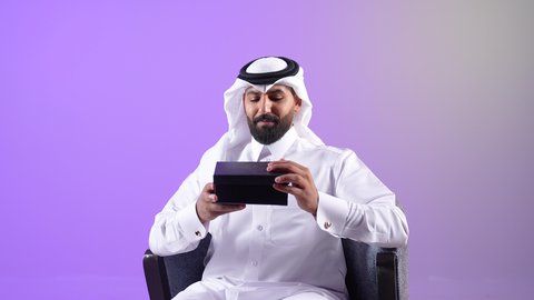 فن انتقاء الهدايا بعناية، بورتريه لرجل عربي خليجي قطري يرتدي الثوب التقليدي والغترة يجلس على كرسي ويقوم بفتح علبة باللون الأسود، مفهوم المفاجأة والإهداء،شاب سعودي يشعر بإيماءات الذهول والسرور بالحصول على هدية ثمينة، خلفية بنفسجية