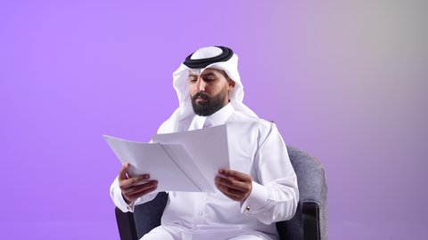 قراءة الملاحظات والمعلومات، بورتريه لرجل عربي خليجي قطري يرتدي الثوب التقليدي والغترة يجلس على كرسي مريح يقوم بتدقيق الاوراق، مفهوم انجاز العمل وتحقيق المهام المطلوبة،شاب سعودي يقوم متابعة الخطط والمشاريع، خلفية بنفسجية