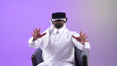 دمج التسلية مع التكنولوجيا والتقنيات الحديثة، الاندماج والتفاعل ومحاكاة العالم الخيالي، بورتريه لرجل عربي خليجي قطري يرتدي الثوب التقليدي والغترة ونظارات الواقع الافتراضي يجلس على كرسي مريح، شاب سعودي يقوم بمد يديه الى الامام بتعابير صد شيء ما والدفاع عن النفس، خلفية بنفسجية