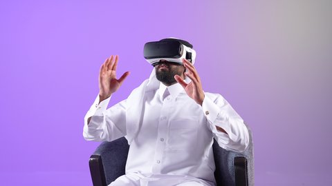 Integration, interaction and simulation of the imaginary world, portrait of a Qatari Gulf Arab man wearing traditional thobe, ghutra and virtual reality glasses sitting on a comfortable chair, a Saudi young man extending his hands forward with expressions of repelling something and self-defense, merging entertainment with technology and modern techniques, purple background