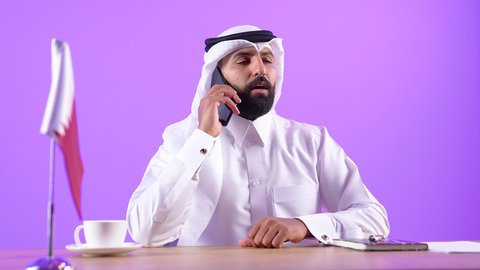 استخدام الهاتف المحمول في العمل، دمج التقنيات والتكنولوجيا بالحياة اليومية، الرد والإجابة على الأسئلة والاستفسارات والمكالمات، بورتريه لرجل عربي خليجي قطري يرتدي الثوب التقليدي والغترة يجلس خلف المكتب يمسك الجوال بيده،خلفية بنفسجية