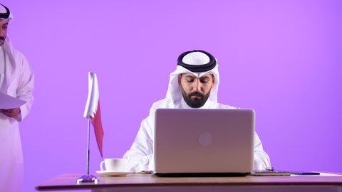 Expressing opinions about strategies and goals, dialogue and exchange of experiences between employees, using a laptop at work, portrait of a Qatari Gulf Arab man wearing a traditional thobe and ghutra sitting behind an office table with the Qatari flag next to him discussing work matters with his colleague, purple background