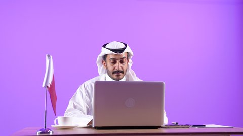 التركيز والتفاني والعمل الجاد، استخدام التقنيات الحديثة في الأعمال الإدارية والمكتبية، التوظيف في المكاتب والشركات القطرية، بورتريه مقرب لرجل عربي خليجي قطري يرتدي الثوب التقليدي والغترة يجلس خلف المكتب وبجانبه علم قطر وأمامه الحاسب المحمول، خلفية بنفسجية