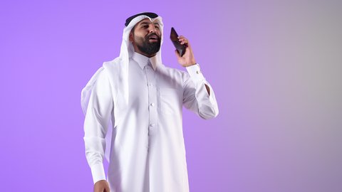استخدام التقنيات والأجهزة الحديثة، شاب سعودي يقوم بالتواصل مع الأهل والأصدقاء، ايماءات السعادة و السرور، بورتريه لرجل عربي خليجي قطري مبتسم يرتدي الثوب التقليدي والغترة يجري مكالمة هاتفية عبر الهاتف المحمول، خلفية بنفسجية