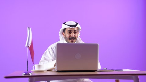 اجراء مكالمة فيديو بايماءات السعادة و السرور، دمج التقنيات الحديثة بالمجال الإداري، العمل في المكاتب والشركات القطرية، الاستمتاع بتناول فنجان من القهوة الساخنة، بورتريه لرجل عربي خليجي قطري يرتدي الثوب التقليدي والغترة يجلس خلف طاولة المكتب وبجانبه علم قطر يعمل على الحاسوب المحمول، خلفية بنفسجية