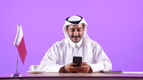 Using a mobile phone at work, holding online meetings via video calls, answering and responding to questions, inquiries and calls, close-up portrait of a Qatari Gulf Arab man wearing a traditional thobe and ghutra sitting behind a desk next to the Qatari flag holding a mobile phone, purple background