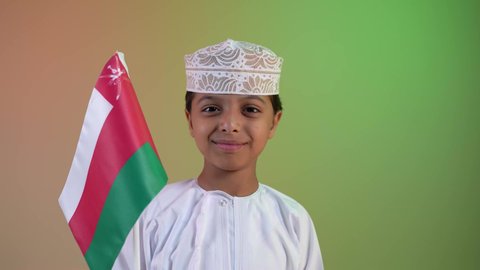 مفهوم حب الوطن والإنتماء إليه، الاعتزاز والفخر بالرموز العمانية، إحياء ذكرى اليوم الوطني العماني، تصوير مقرب لولد عربي خليجي عماني يرتدي الدشداشة والكمه يمسك علم لسلطنة عمان ينظر الى العلم بايماءات الفرح والسرور